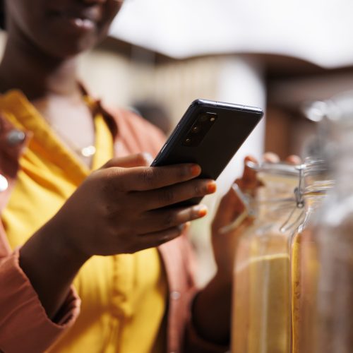 Close-up shot of black woman using cell phone to search for recipes and compare prices at local store. African American female creates modern and convenient shopping experience with use of smartphone.
